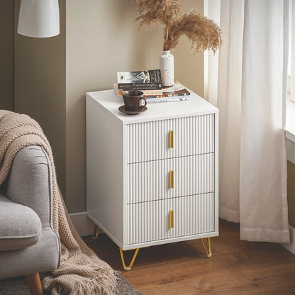 Table de chevet blanche à trois tiroirs près d’un fauteuil gris, avec un vase et un livre sur le plateau