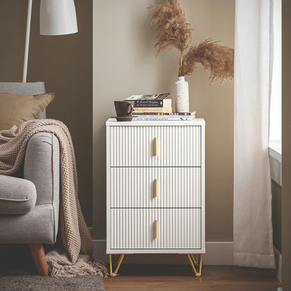 Table de chevet blanche à trois tiroirs près d’un canapé beige, dans une pièce lumineuse avec moulures et parquet clair