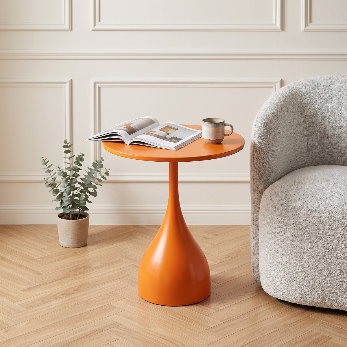 Une photo de style magazine de décoration d'une table d'appoint tulip-base orange brillant dans un salon moderne et chaleureux. Elle est posée sur un parquet en chevrons clair, à côté d'un fauteuil texturé. Sur la table, un livre d'art et une tasse de café. Le fond montre des moulures murales subtiles et un mur blanc chaud. Éclairage naturel doux