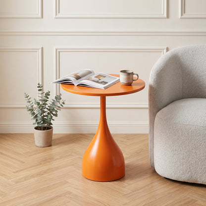 Une photo de style magazine de décoration d'une table d'appoint tulip-base orange brillant dans un salon moderne et chaleureux. Elle est posée sur un parquet en chevrons clair, à côté d'un fauteuil texturé. Sur la table, un livre d'art et une tasse de café. Le fond montre des moulures murales subtiles et un mur blanc chaud. Éclairage naturel doux
