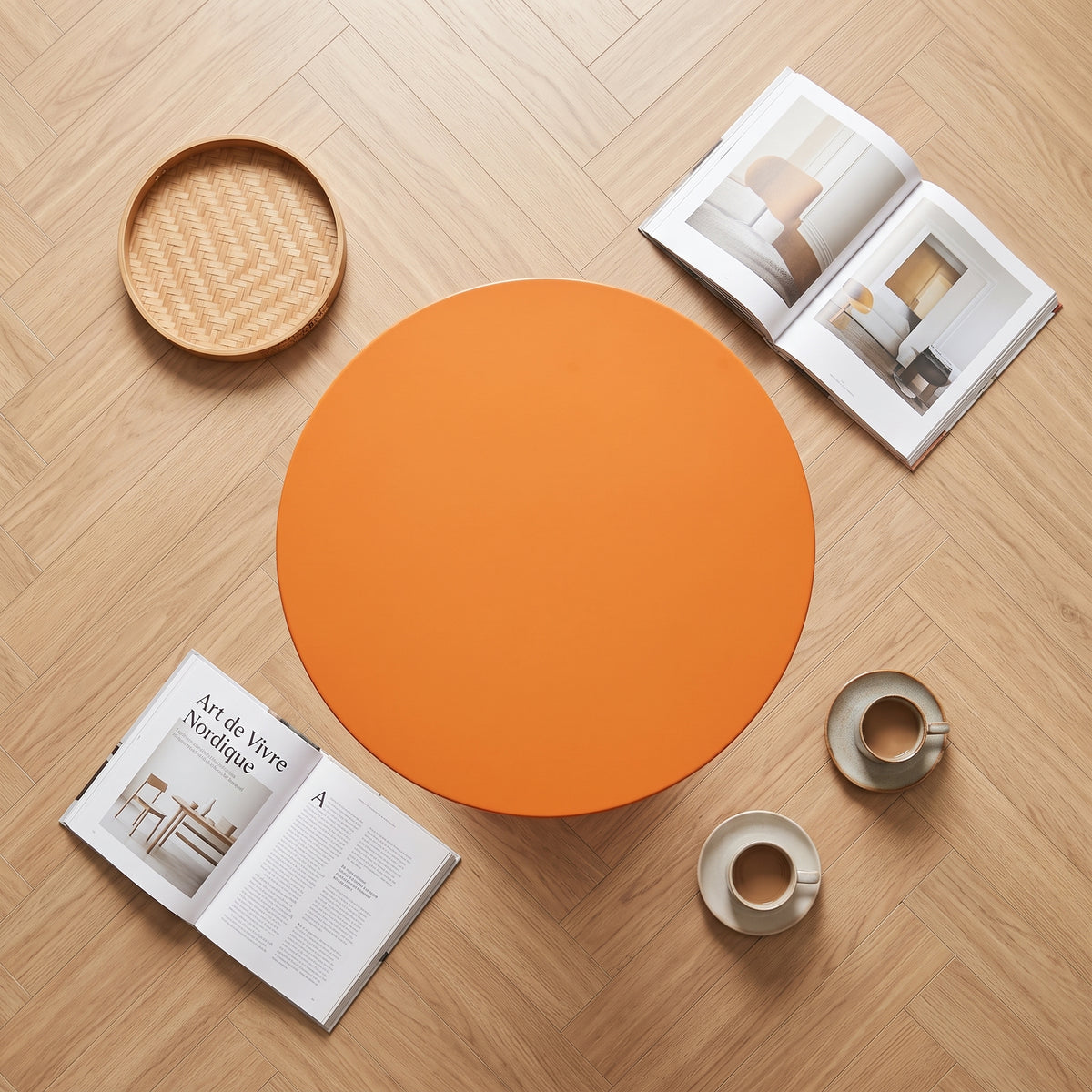 Une photo carrée de style magazine de décoration en vue de dessus (flat lay) de la table d'appoint tulip-base orange. La table est centrée sur le parquet en chevrons clair. Autour d'elle sont disposés un plateau en rotin tissé, un livre ouvert intitulé 'Art de Vivre Nordique', et deux tasses à café minimales avec soucoupes. L'ensemble est curated, lumineux et stylé.